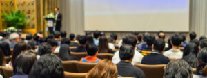 A large group of people in a conference room with a presenter at a podium