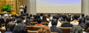 A large group of people in a conference room with a presenter at a podium