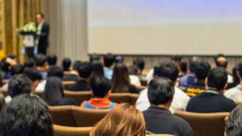 A large group of people in a conference room with a presenter at a podium