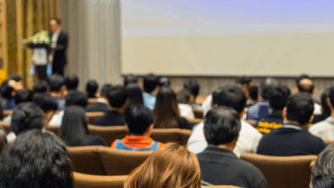 A large group of people in a conference room with a presenter at a podium