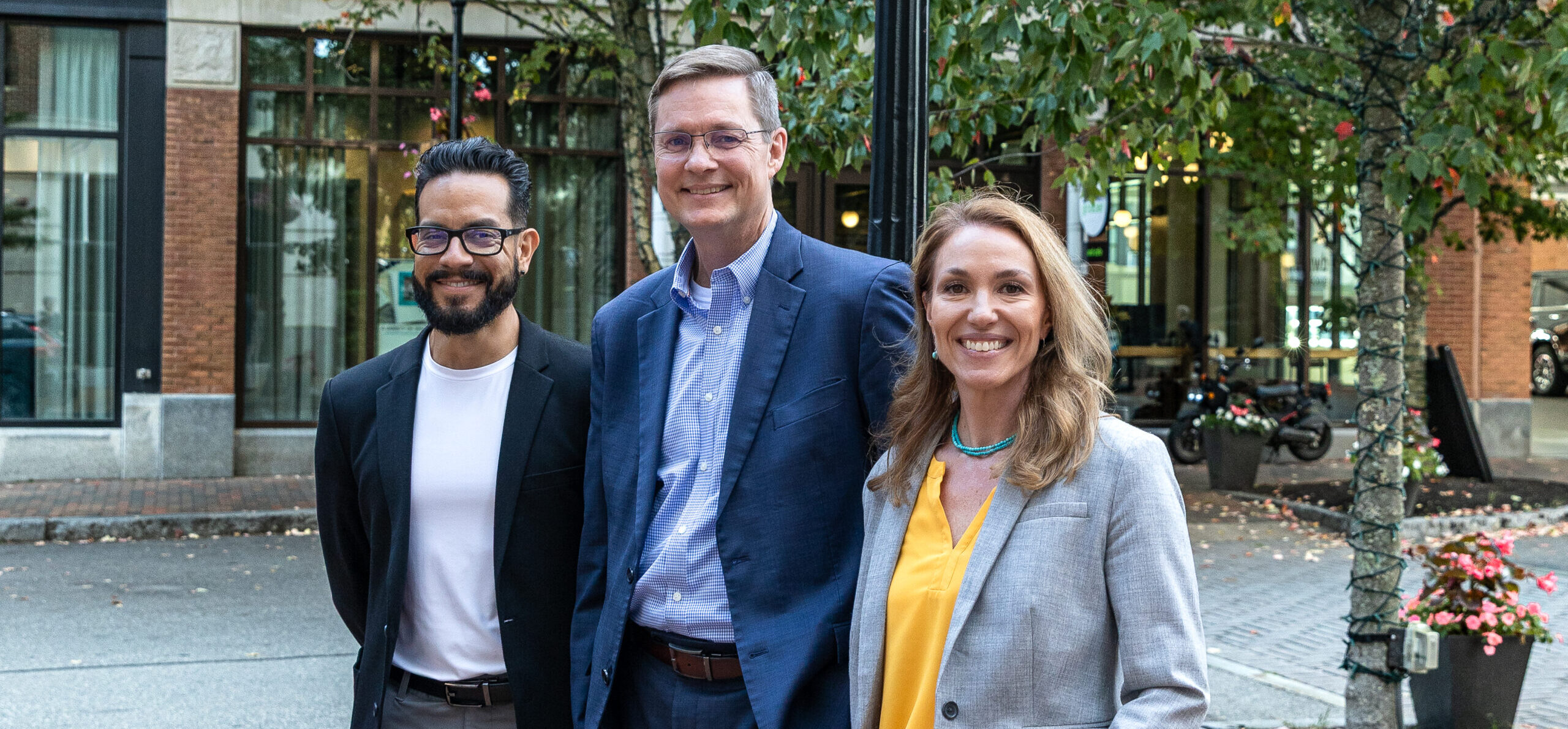 Three people standing side by side, smiling at the camera with trees and a building in the background.