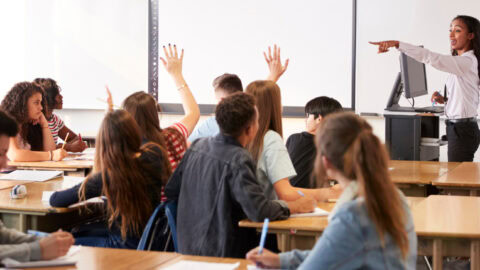 A classroom of students with their hands raised, sitting at desks.