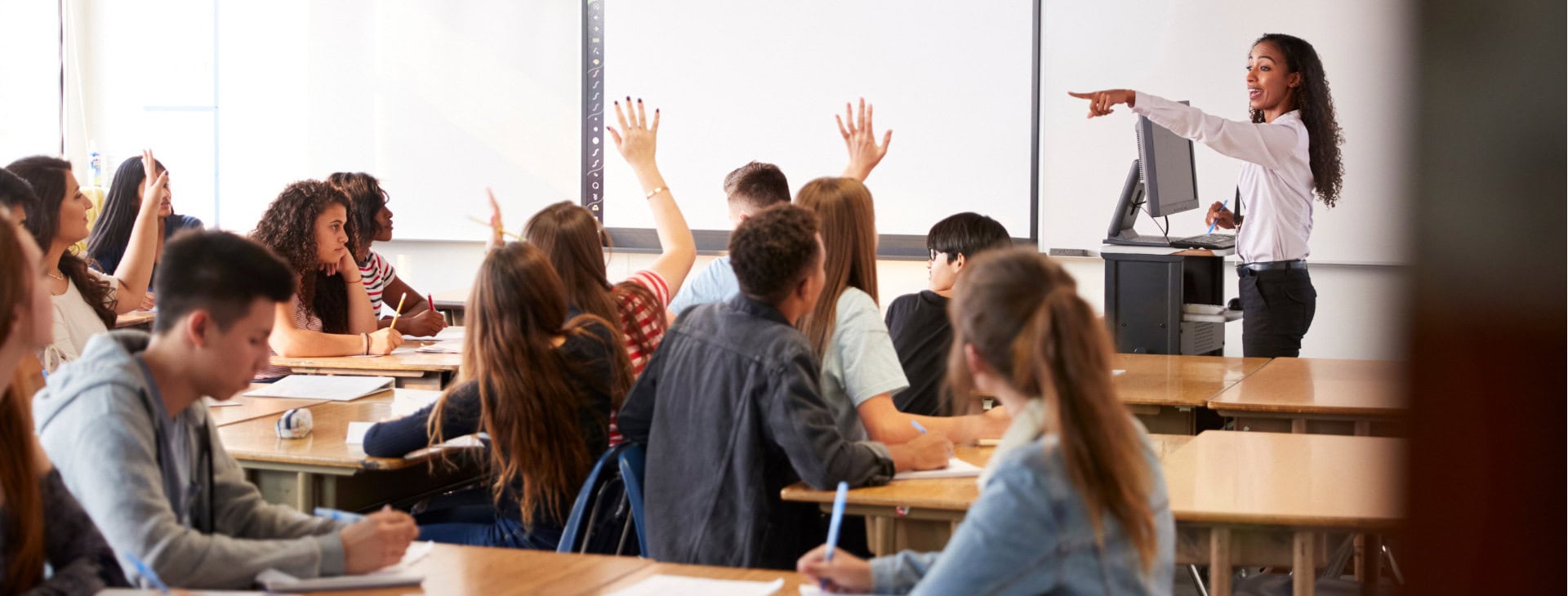 A classroom of students with their hands raised, sitting at desks.