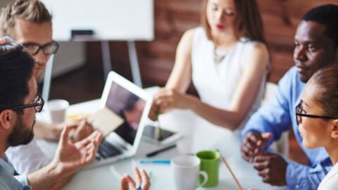 A group of professionals in a meeting, sitting at a table with laptops.