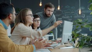 Four people sitting at a table have a discussion while looking at a desktop computer screen.