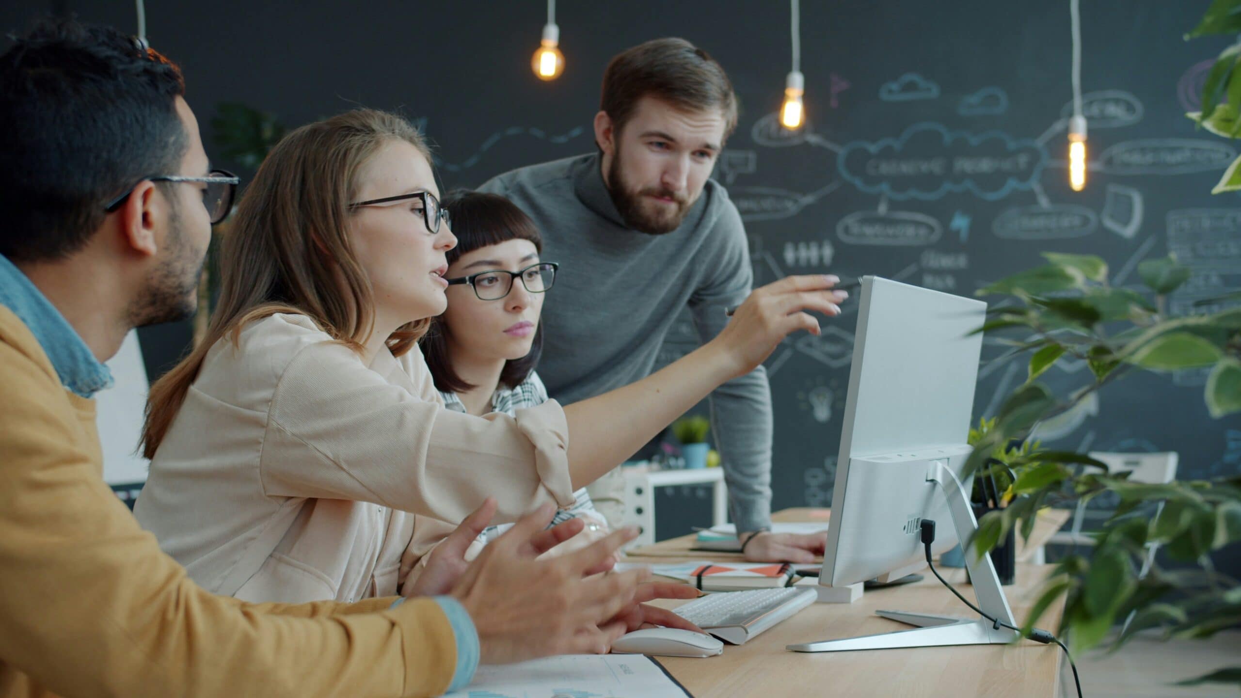 Four people sitting at a table have a discussion while looking at a desktop computer screen.